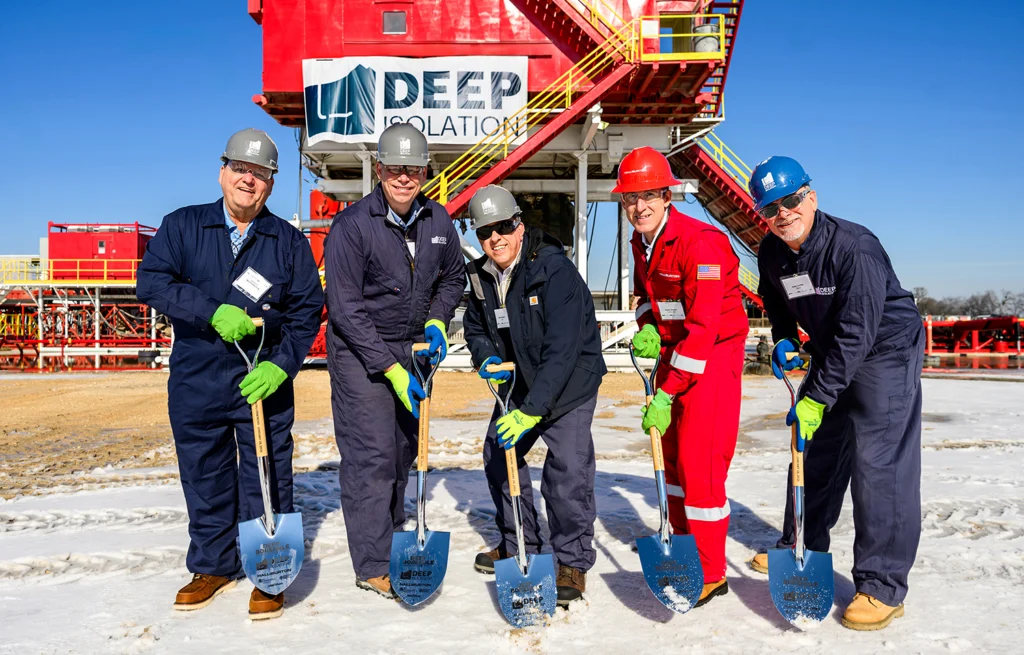 Team of executives pose with shovels in front of red drill rig. Deep Isolation banner is visible on the rig.